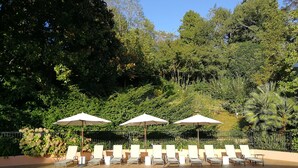 Un área junto a la piscina con sillones de descanso y sombrillas, rodeada de exuberante vegetación y árboles.