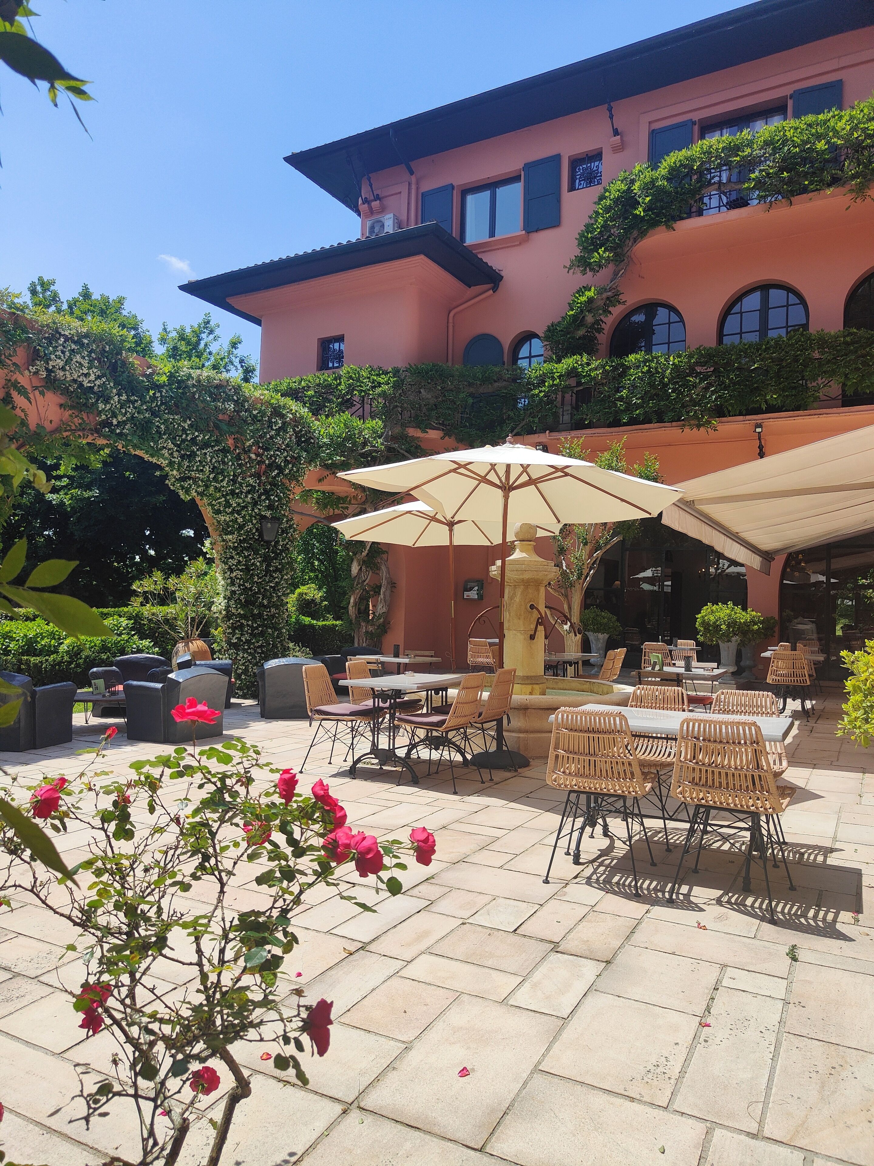 Un patio al aire libre con mesas y sillas, una fuente y un edificio con ventanas de arco en el fondo.