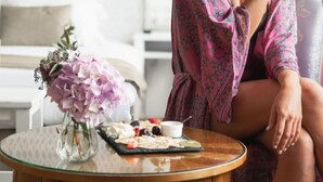 Una mujer sentada en una silla en una habitación con una cama, una mesa redonda con un jarrón de flores y una bandeja de comida.