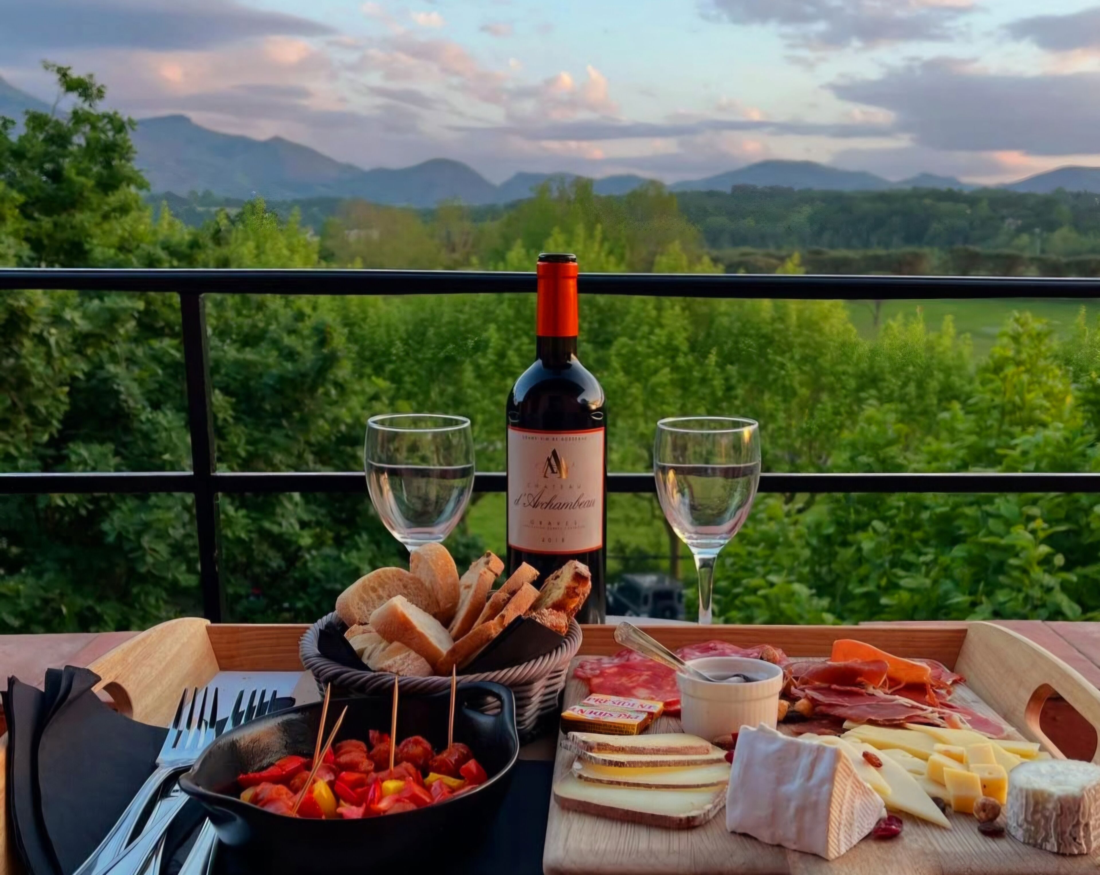 Une table avec une bouteille de vin, des verres, un panier de pain, une planche de charcuterie et un saladier rempli de brochettes de fruits.