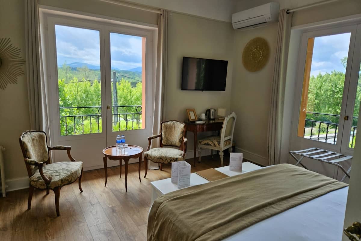 Habitación de hotel con cama, dos sillones, una mesita con una botella de agua, televisor en la pared y balcón con vistas al verdor.