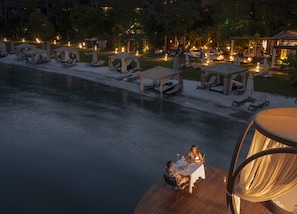 A lakeside dining area with tables and chairs, surrounded by lounge chairs and umbrellas, and a wooden deck overlooking the water.
