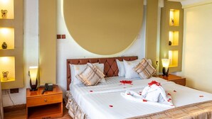 Een keurig opgemaakt bed met dekbed, kussens en een handdoek. Er staat een nachtkastje met een lamp en een telefoon. De kamer heeft een gebogen muur en een plank met decoratieve objecten.