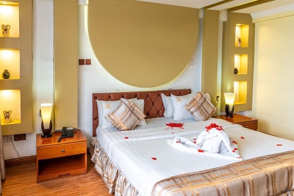 A neatly made bed with a duvet, pillows, and a towel arranged on it. There is a bedside table with a lamp and a phone. The room has a curved wall and a shelf with decorative items.