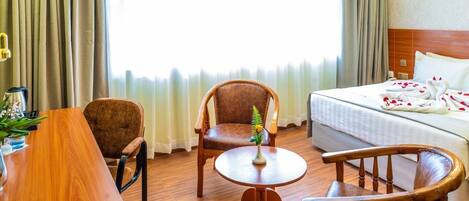 A hotel room with a bed, a desk, two chairs, a small table with a vase, and curtains.