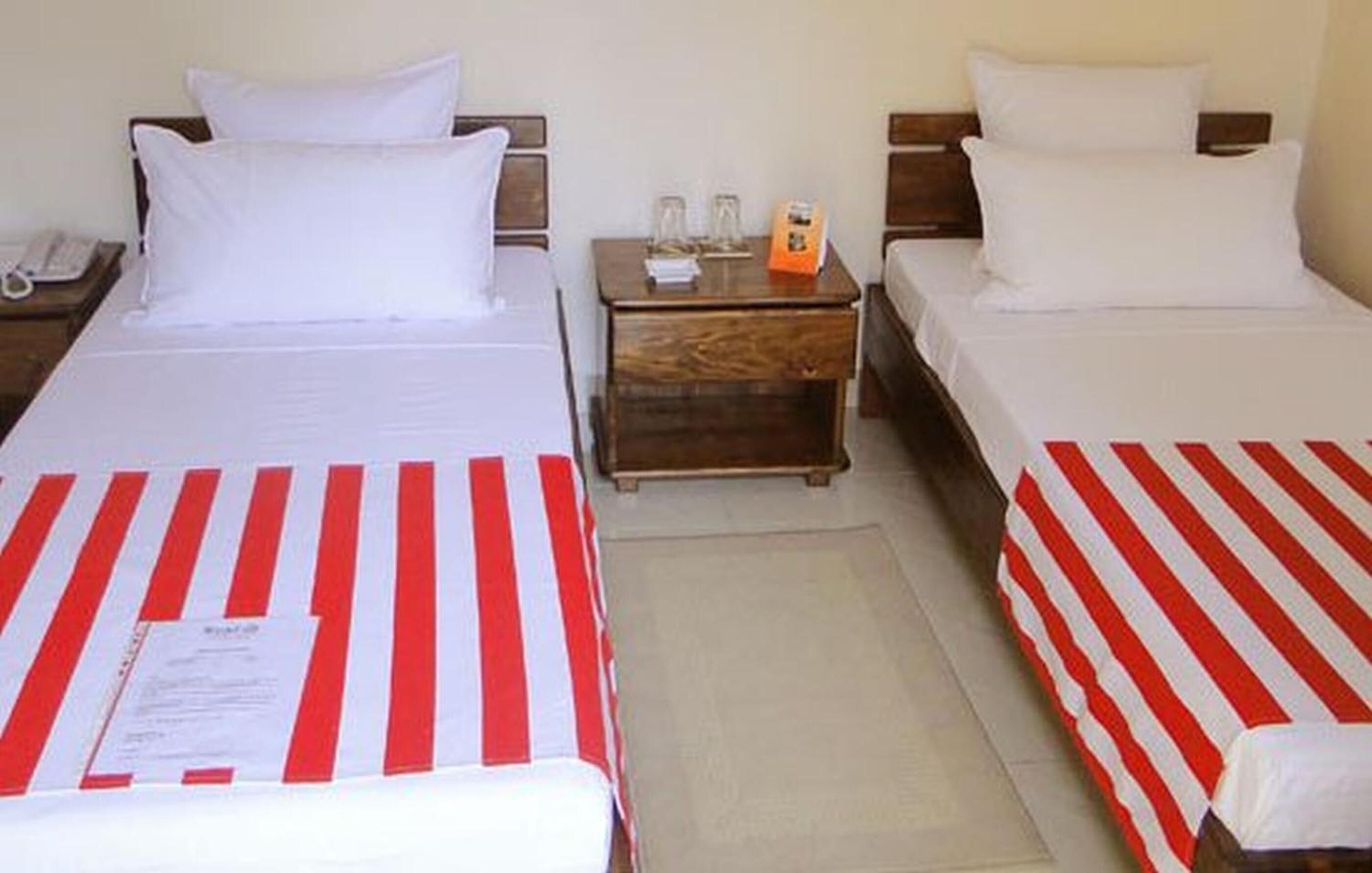 A hotel room with two beds, each with white bedding and red-striped bedspreads. There is a wooden nightstand between the beds with a telephone and a small table with a glass and a book.