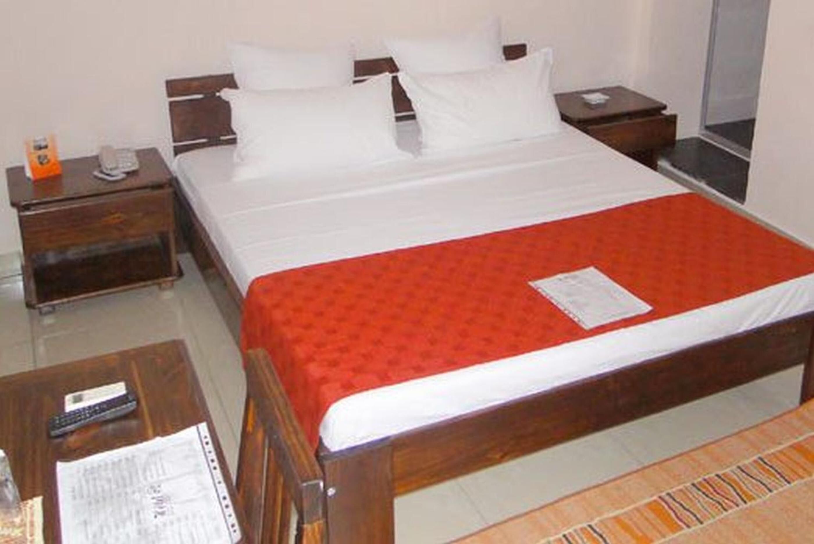 A hotel room with a wooden bed, white bedding, a red blanket, a nightstand with a phone, and a small table with a remote control and papers.