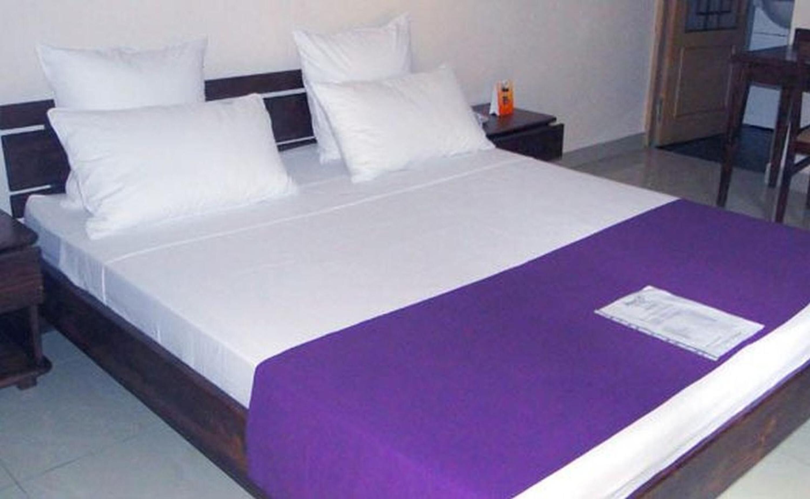 A neatly made bed with white linens and a purple blanket, a wooden nightstand with a bottle, and a wooden chair in a simple room.