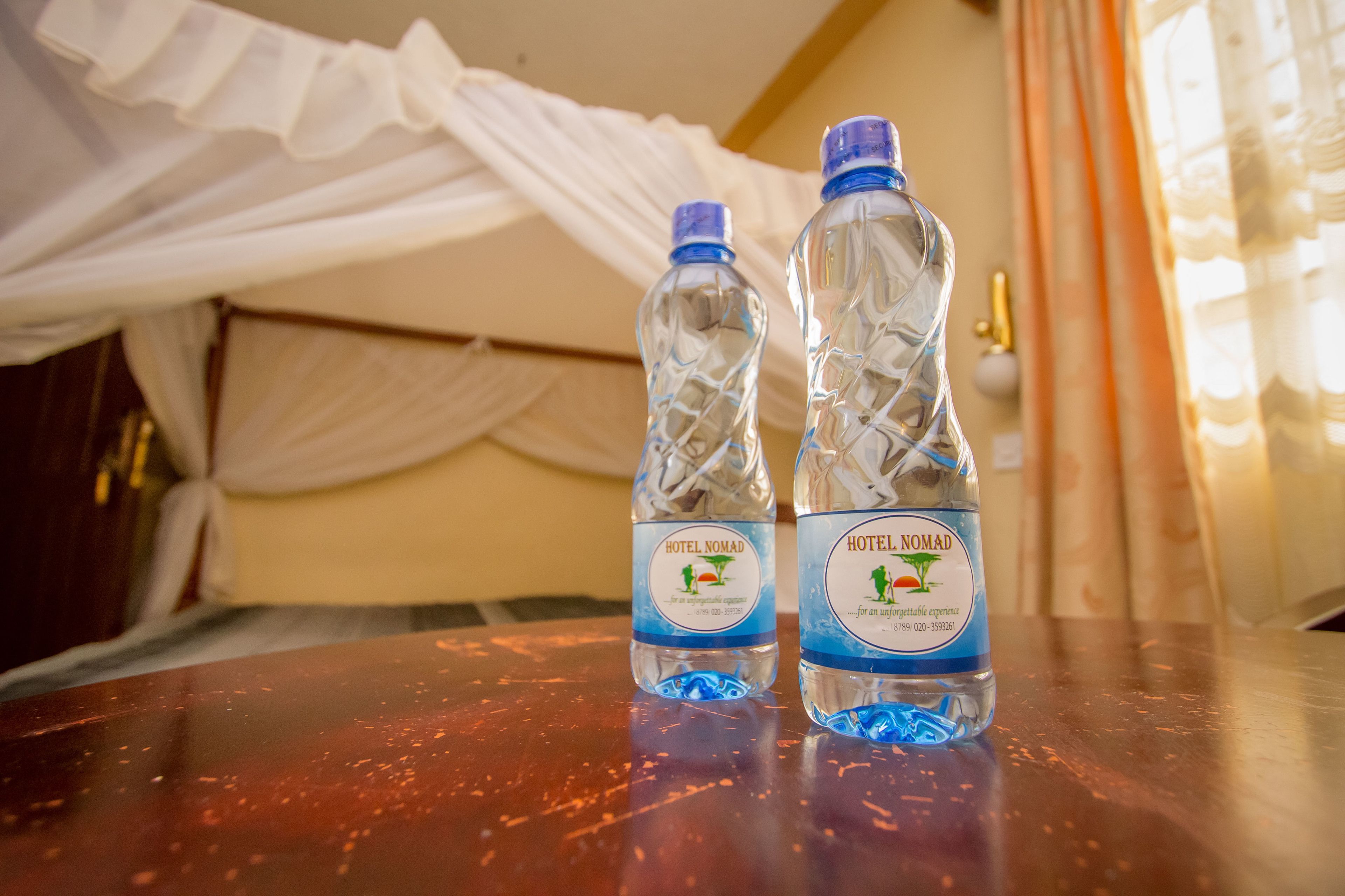 Two Hotel Nomad water bottles on a wooden table in a room with a bed and curtains.
