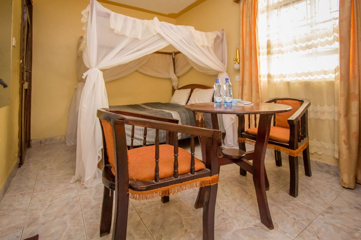 A room with a bed covered by a canopy, two wooden chairs with orange cushions, a small round table with two water bottles, and a window with curtains.