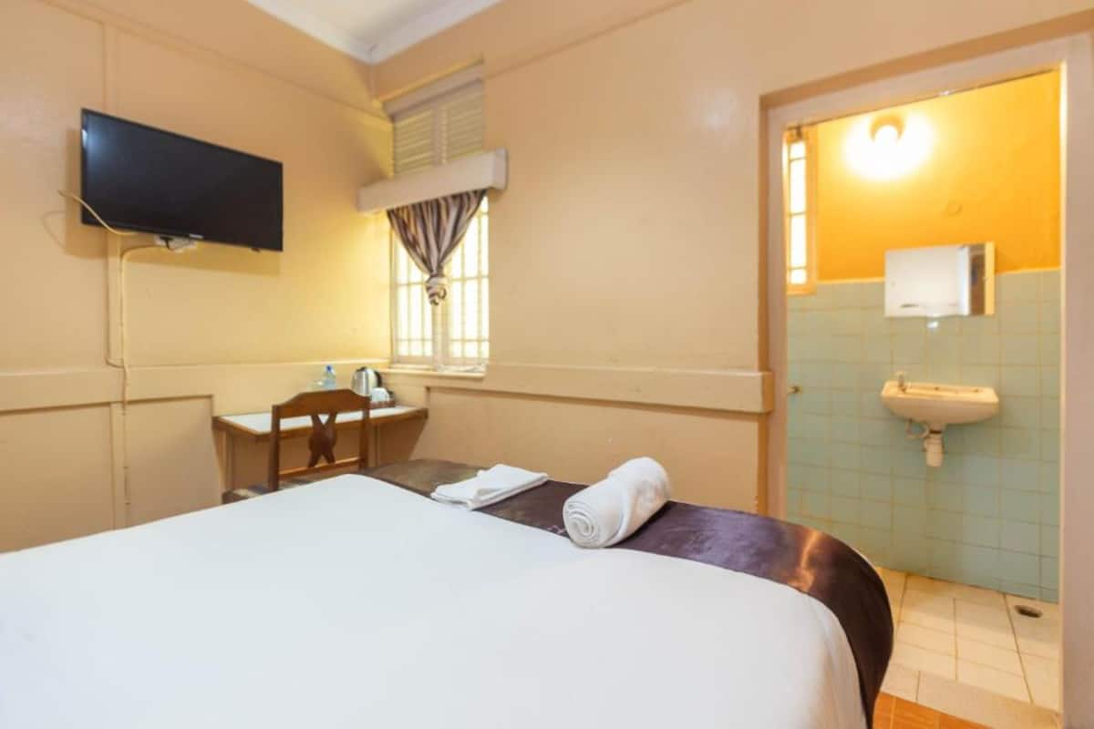 Habitación de hotel con cama, escritorio, silla, televisor, ventana con cortinas y baño con lavamanos y espejo.