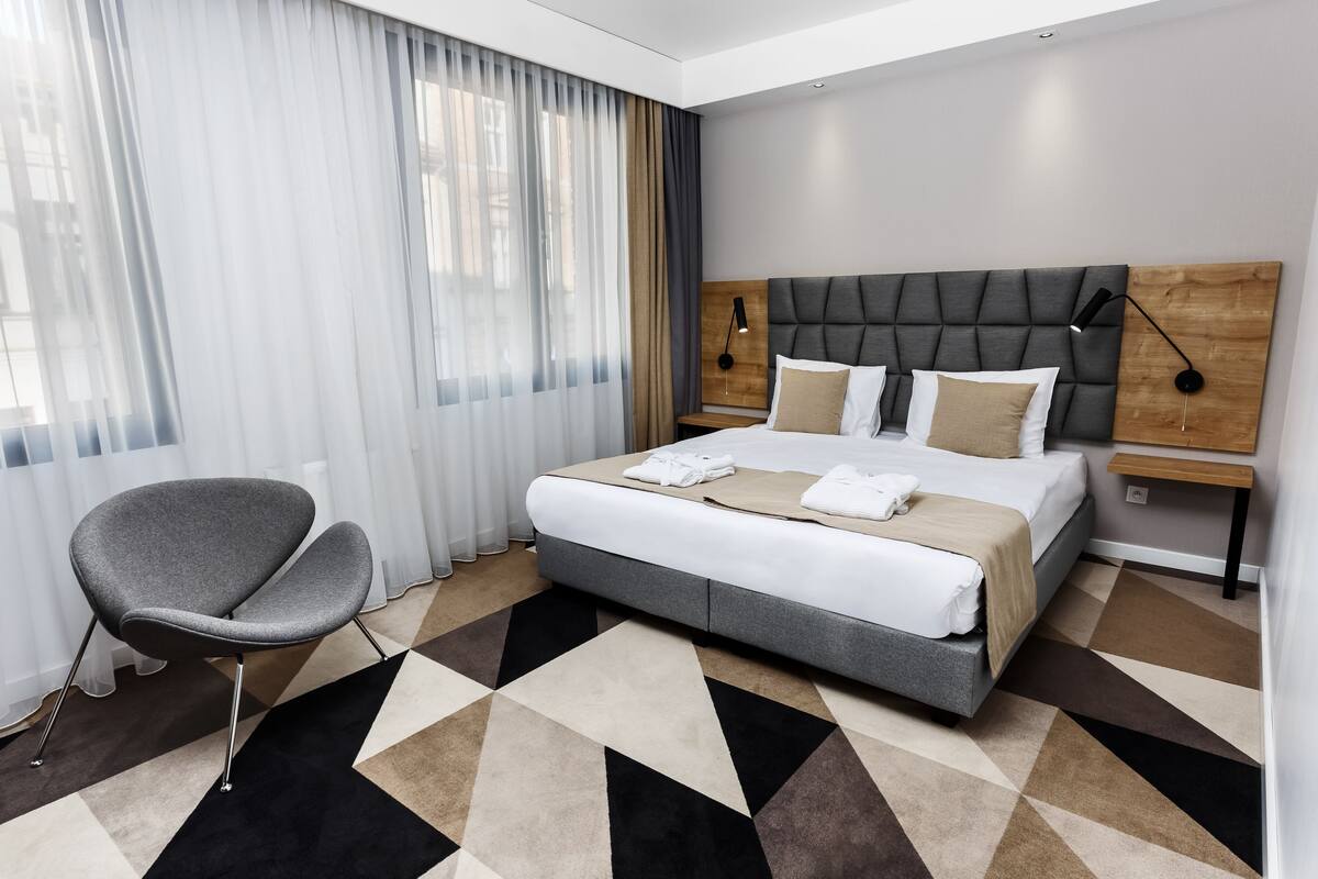 Moderni hotellihuone, jossa on suuri sänky, harmaa nojatuoli ja geometrisesti kuvioitu matto.