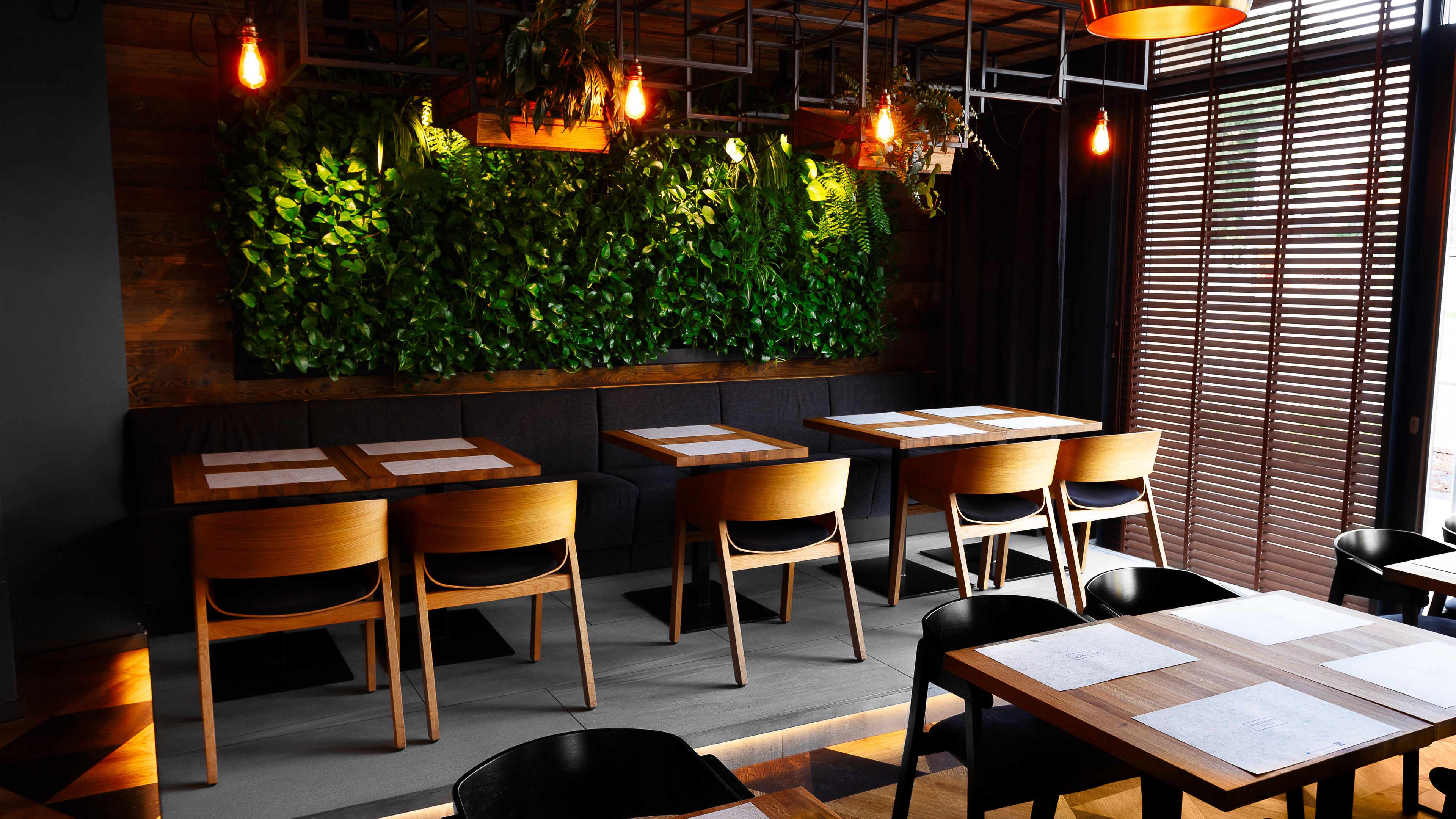 A restaurant interior with wooden tables and chairs, a green vertical garden wall, and hanging light fixtures.