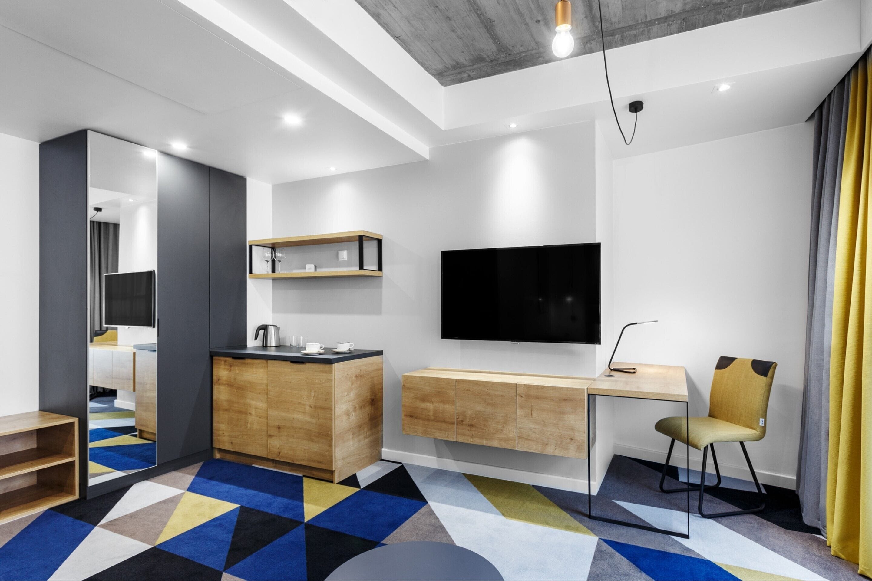 A modern hotel room with a wooden desk, a flat-screen TV, a built-in kitchen, and a colorful geometric carpet.
