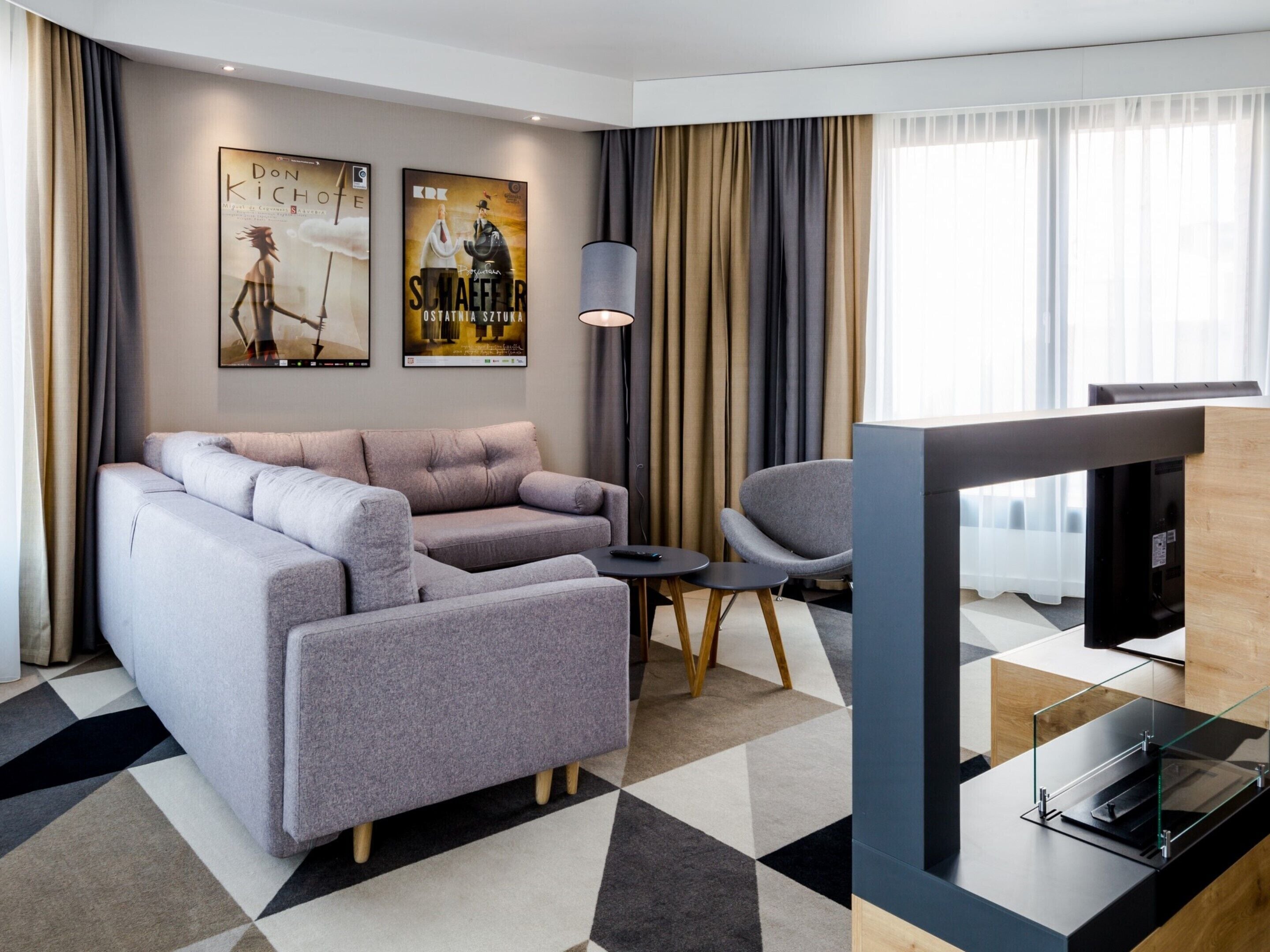 A modern hotel room with a grey sofa, a small table, a chair, and a TV.