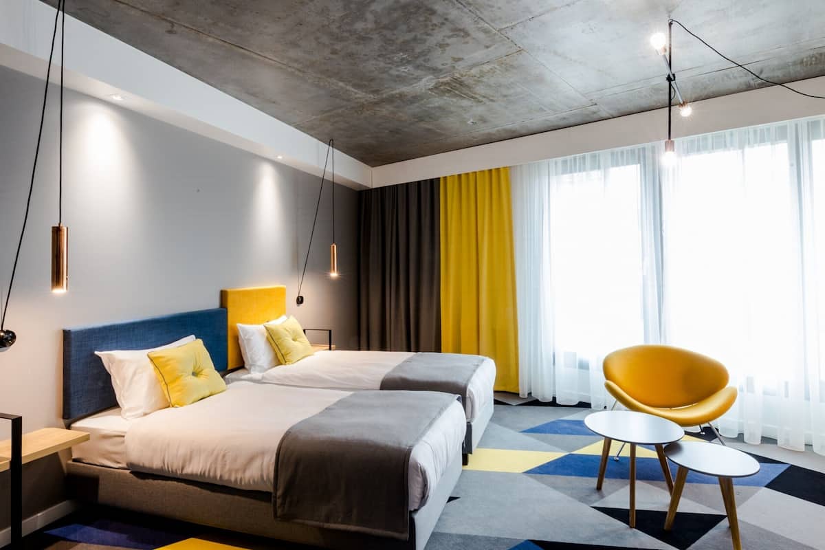 Moderni hotellihuone, jossa on kaksi sänkyä, keltainen tuoli ja geometrisesti kuvioitu matto.
