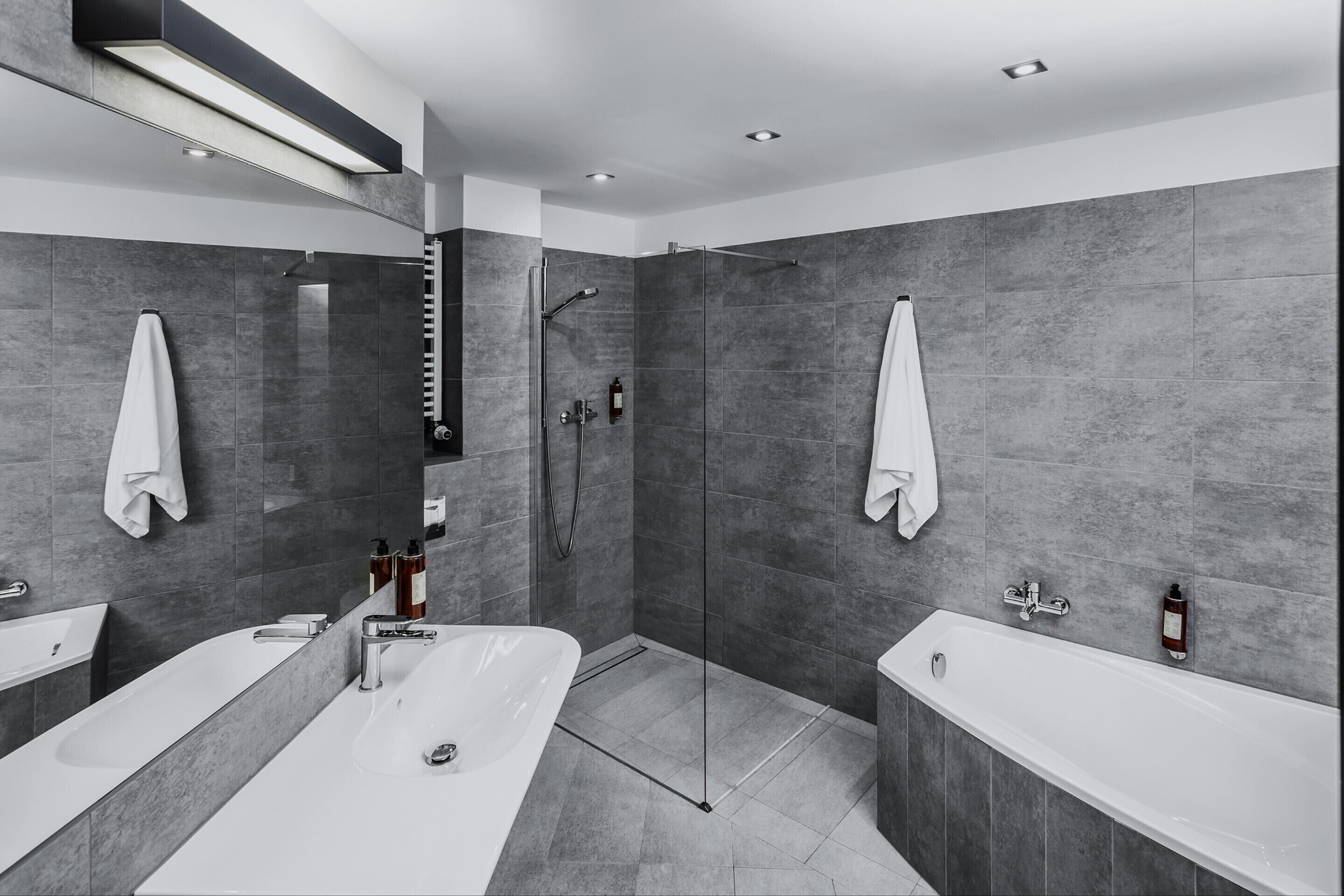 A modern bathroom with two large white tubs, a glass-enclosed shower, and grey tiled walls.
