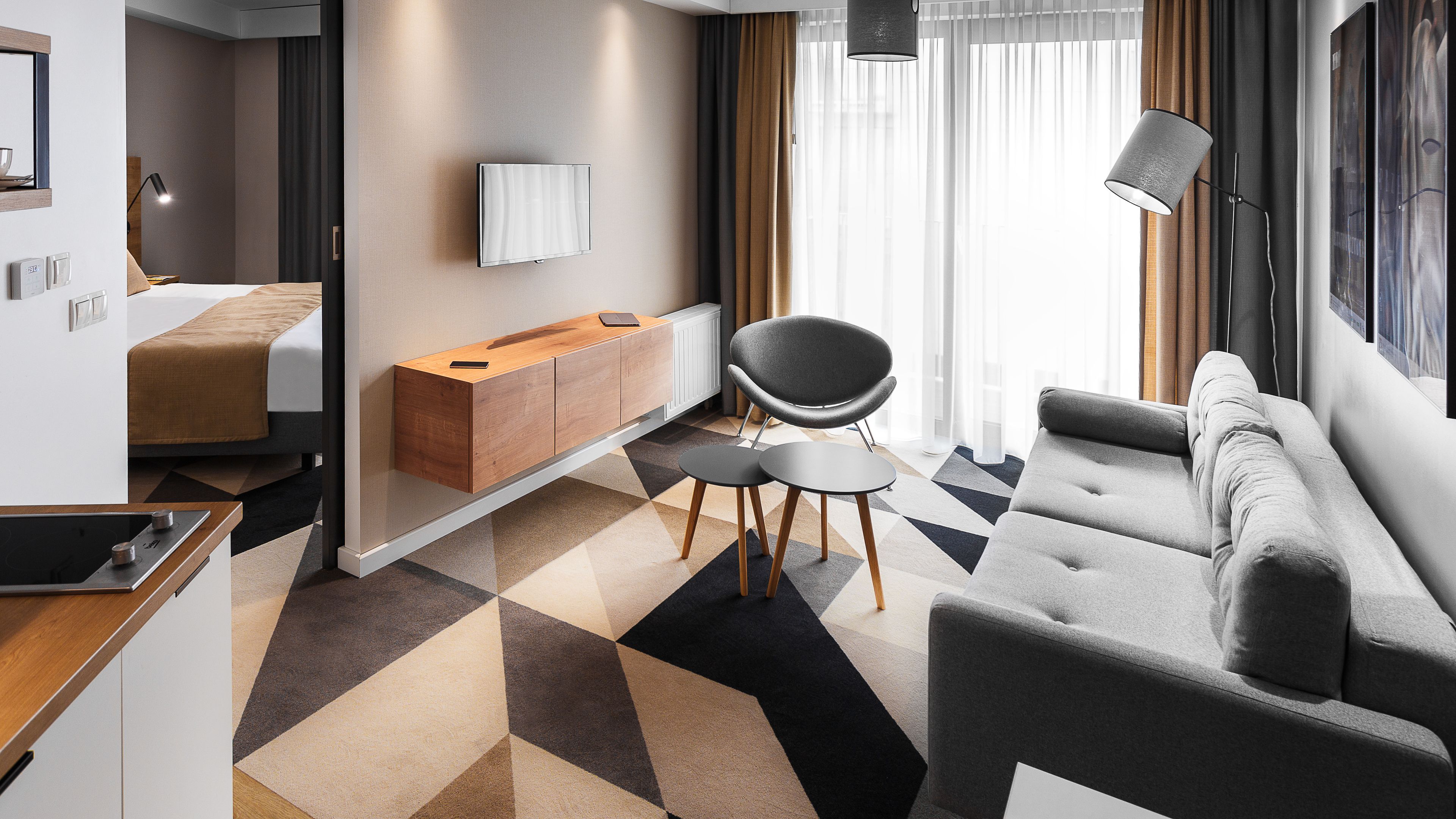 Una habitación de hotel moderna con un sofá, una silla, una mesita, un televisor montado en la pared y un armario de madera.