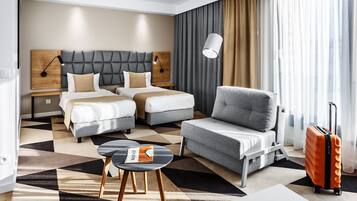 Hotellihuone, jossa on kaksi sänkyä, harmaa nojatuoli, pieni pöytä ja matkalaukku.