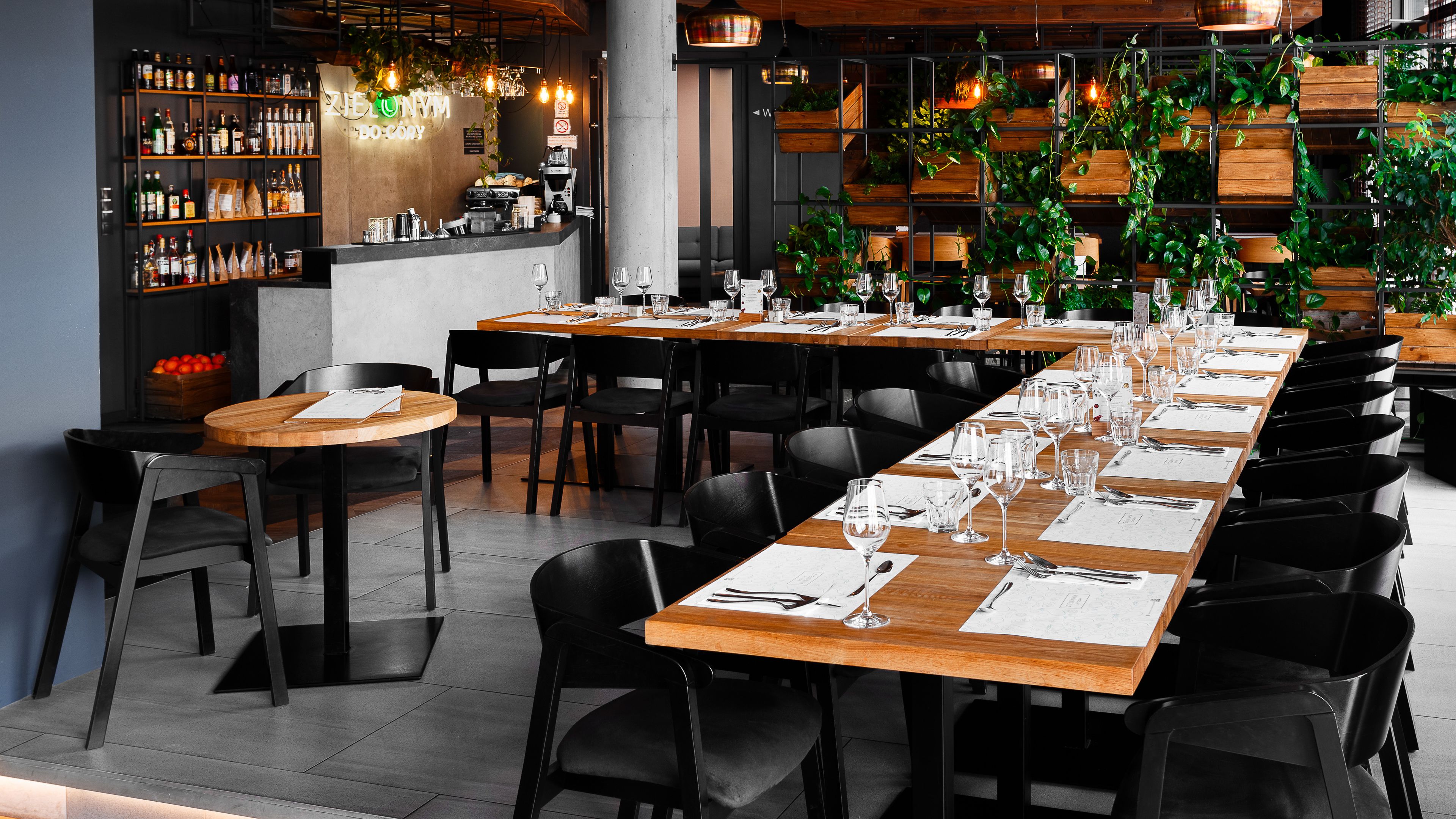 A restaurant with multiple tables set for dining, a bar area with various bottles, and a green wall with plants.