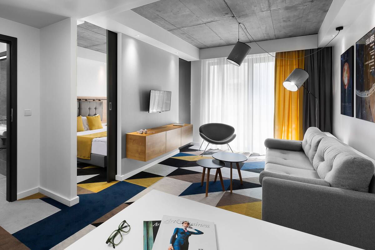 Moderni hotellihuone, jossa on sohva, ruokapöytä, televisio ja kylpyhuone, joka näkyy avoimesta ovesta.