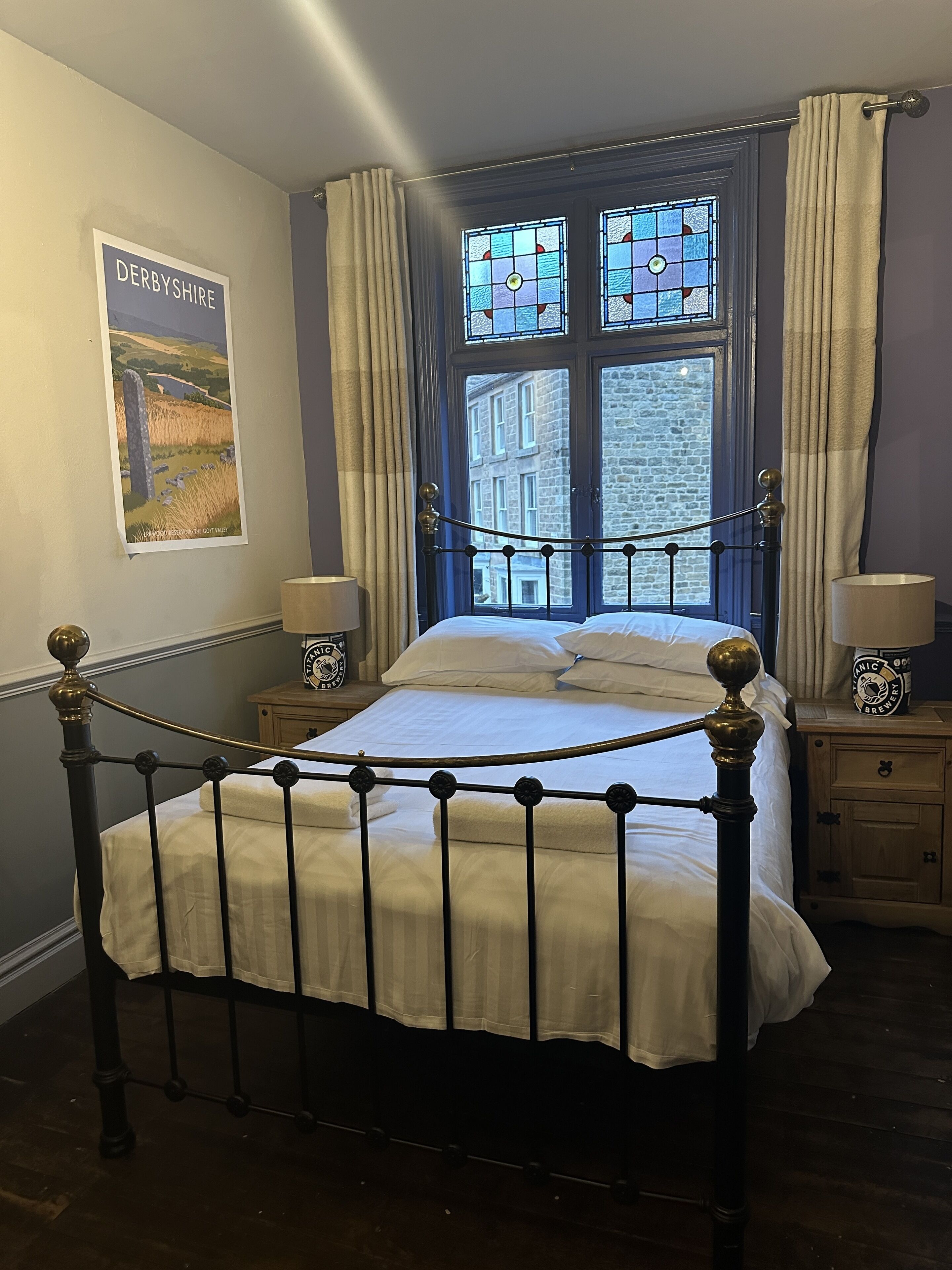 A bedroom with a four-poster bed, a poster of Derbyshire on the wall, and a view of a stone building through a window.