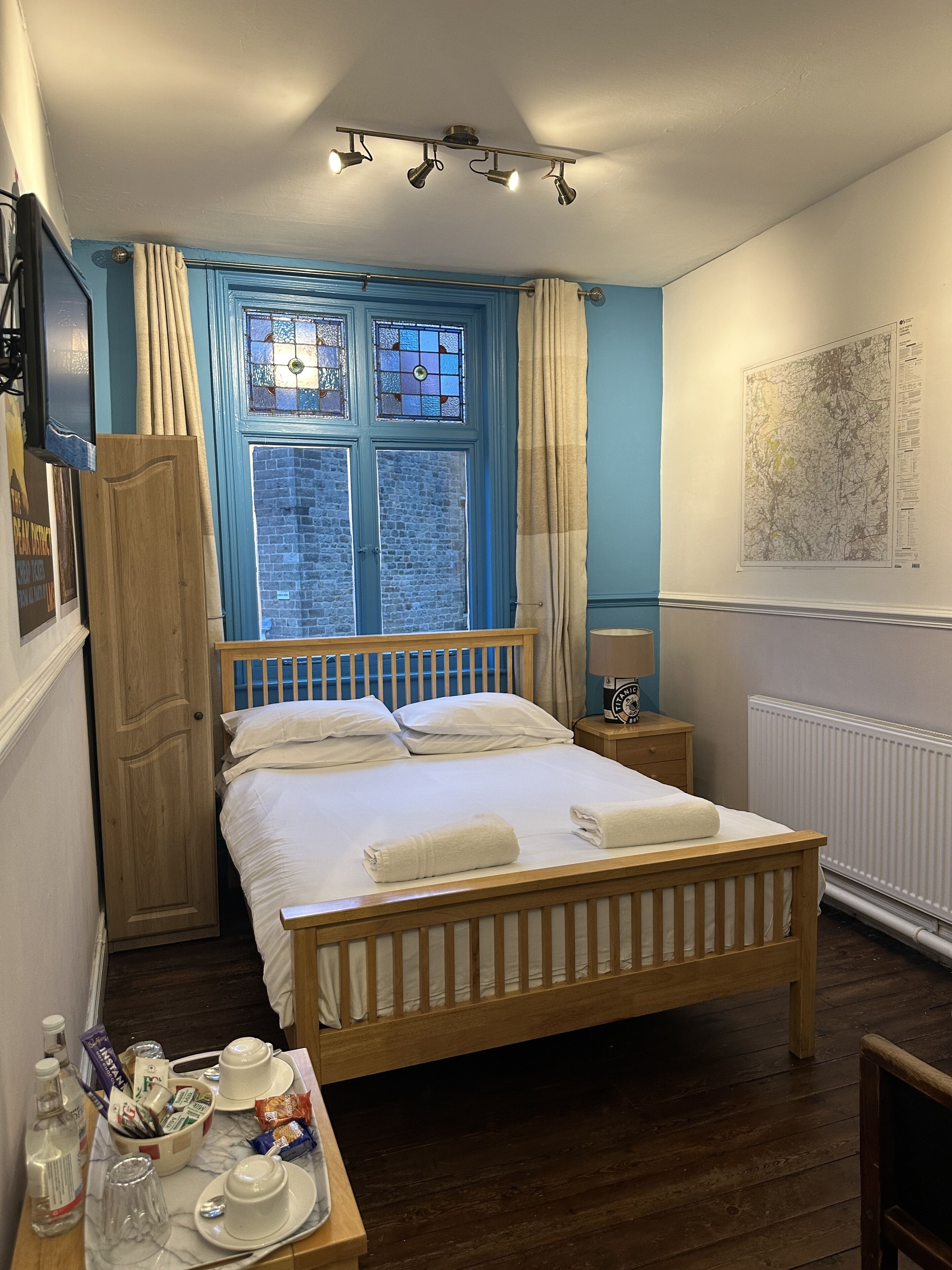 A bedroom with a wooden bed, a nightstand with a lamp, a window with curtains, a map on the wall, and a wooden wardrobe.