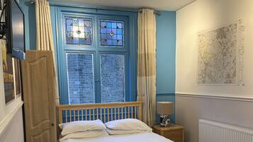 A bedroom with a wooden bed, a nightstand with a lamp, a window with curtains, a map on the wall, and a wooden wardrobe.