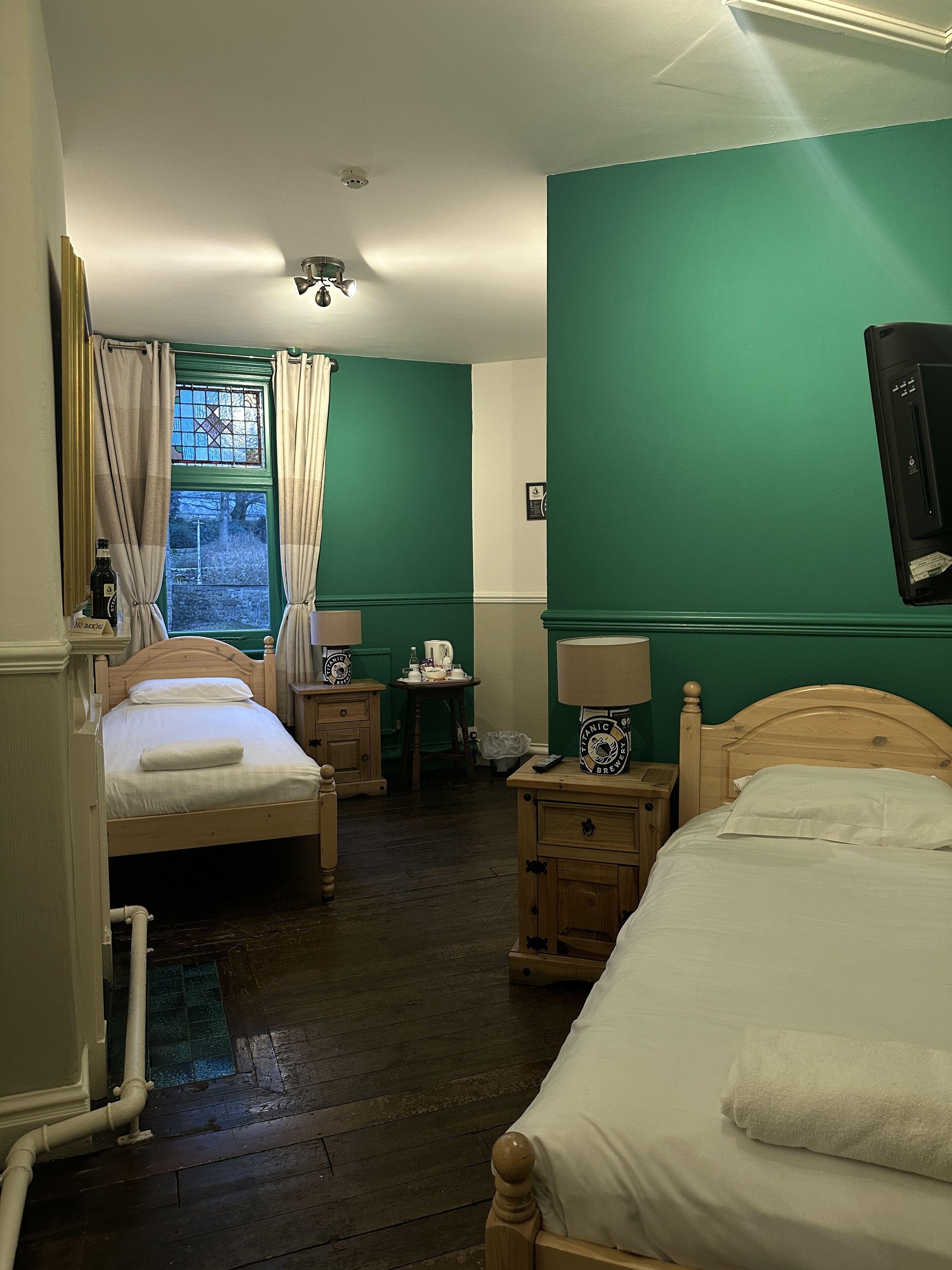 A hotel room with two beds, a window with curtains, a small table with a lamp, and a wall-mounted TV.