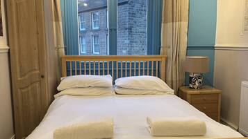 Una cama con ropa de cama y toallas blancas, un cabecero de madera, una mesita de noche con una lámpara y una ventana con cortinas azules.