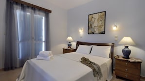 A neatly made bed with white linens, a leopard print throw, and a set of towels on the side. There are two bedside tables with lamps, a wall-mounted painting, and a window with curtains. - Villa Jazz Rock (Corfu)