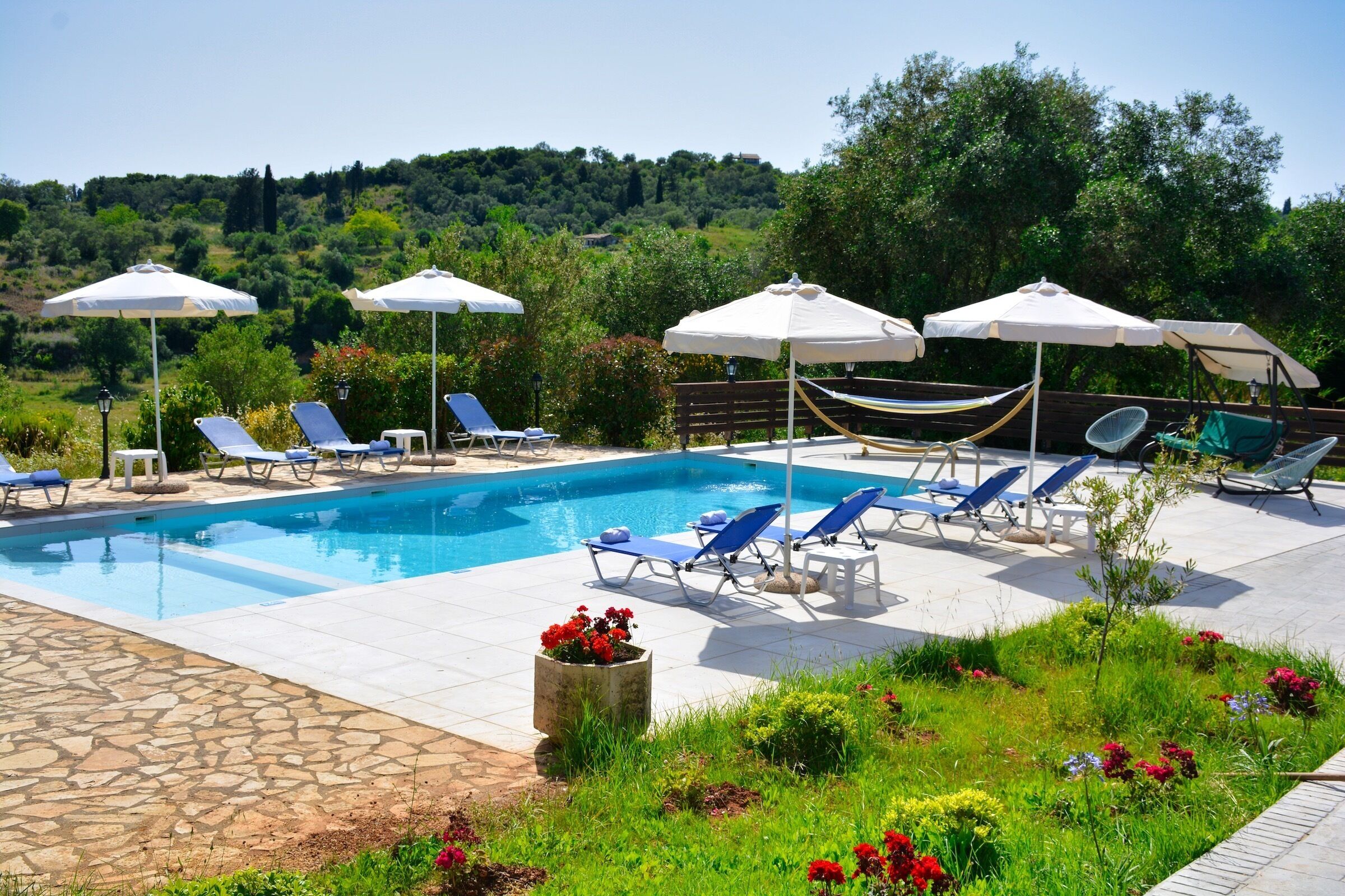 A pool area with lounge chairs and umbrellas, surrounded by greenery and a stone pathway.