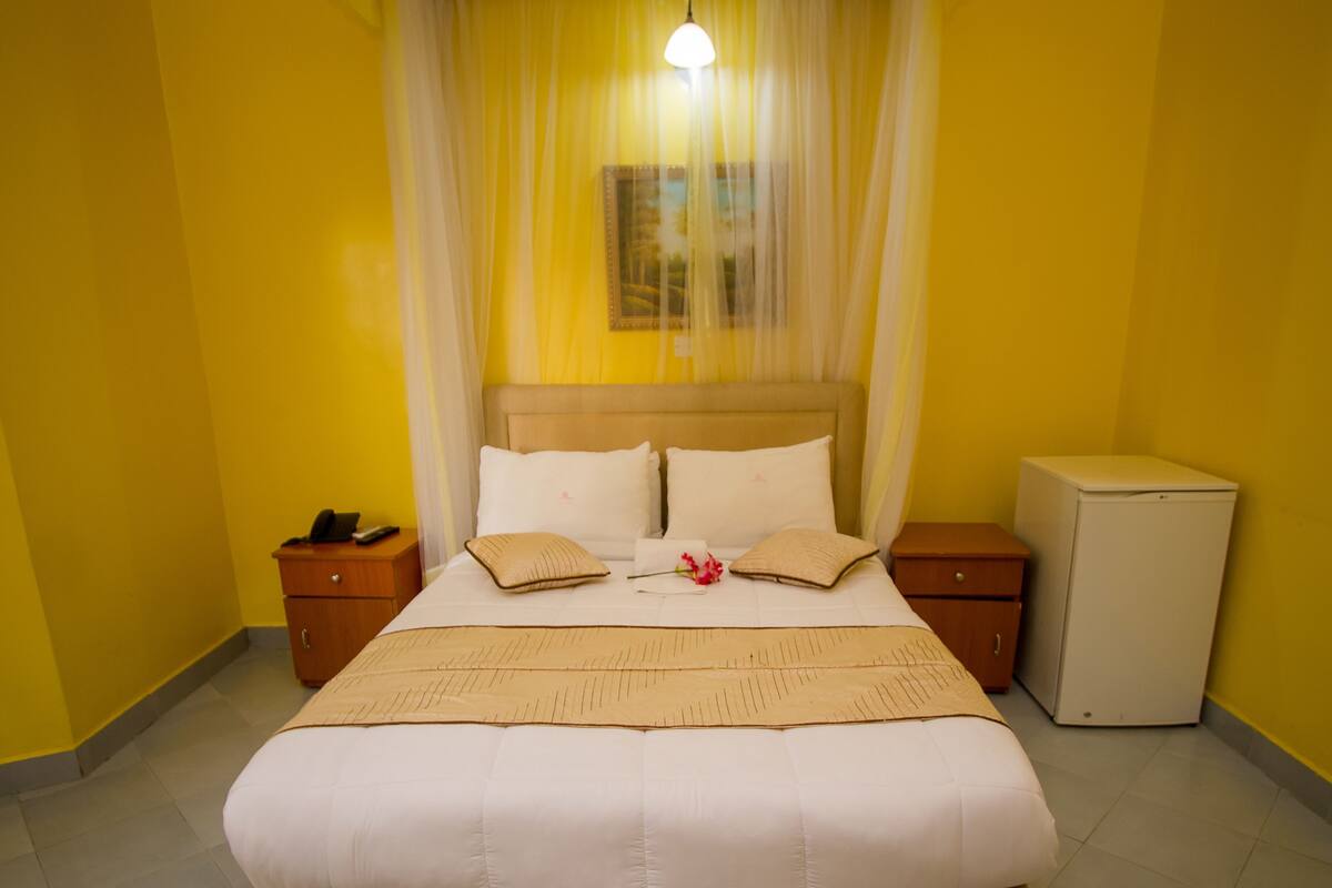 Quarto de hotel com uma cama, mesas de cabeceira, um pequeno frigorífico e uma cortina.
