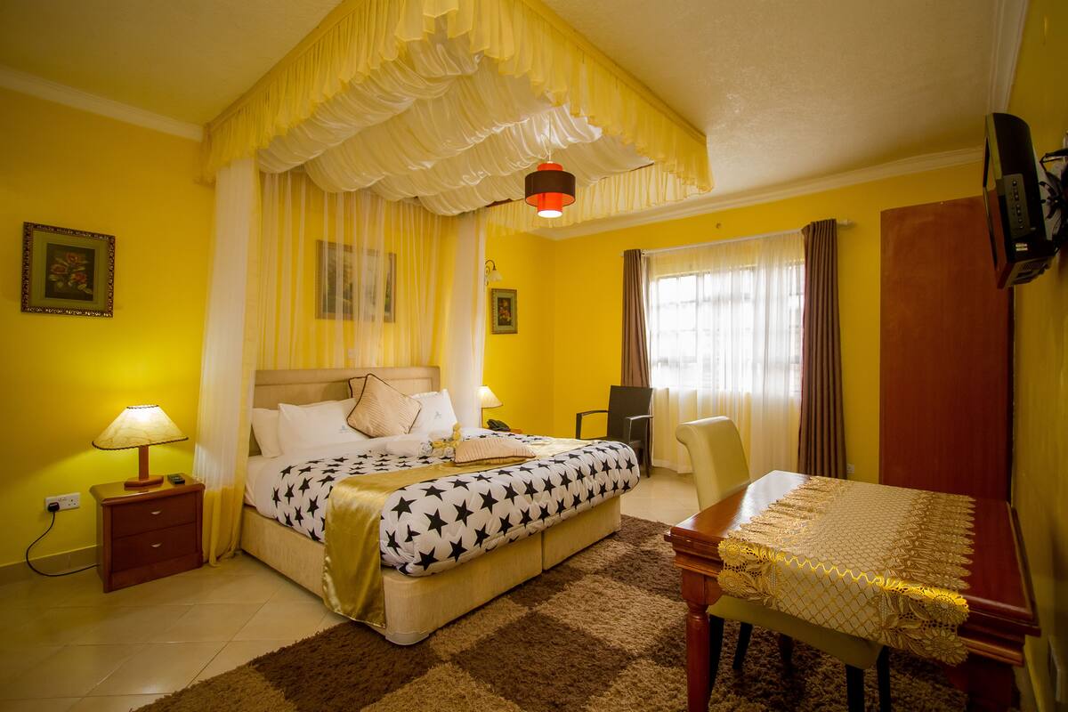 Quarto de hotel com cama, mesa de cabeceira, luminária e uma mesa coberta por um tecido.
