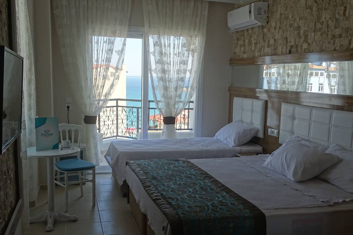 A hotel room with two beds, a desk, and a chair. There is a balcony with a view of the sea.