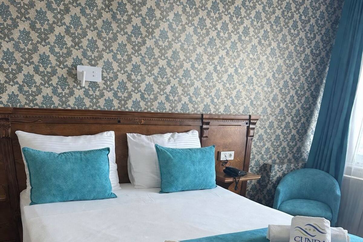 A hotel room with a large bed, two pillows, a bedside table, a chair, and a window with curtains.