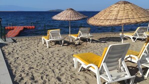 A beach with white lounge chairs and yellow cushions, some under woven umbrellas, and a clear blue sky. - Cunda Basel hotel (Ayvalik)