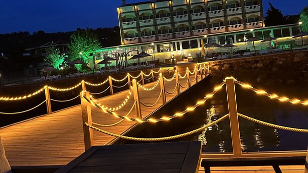 A wooden deck with a rope barrier, outdoor seating, and a building with balconies in the background. - Cunda Basel hotel (Ayvalik)
