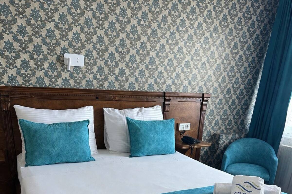A hotel room with a large bed, two pillows, a bedside table, a chair, and a wall with a patterned wallpaper.
