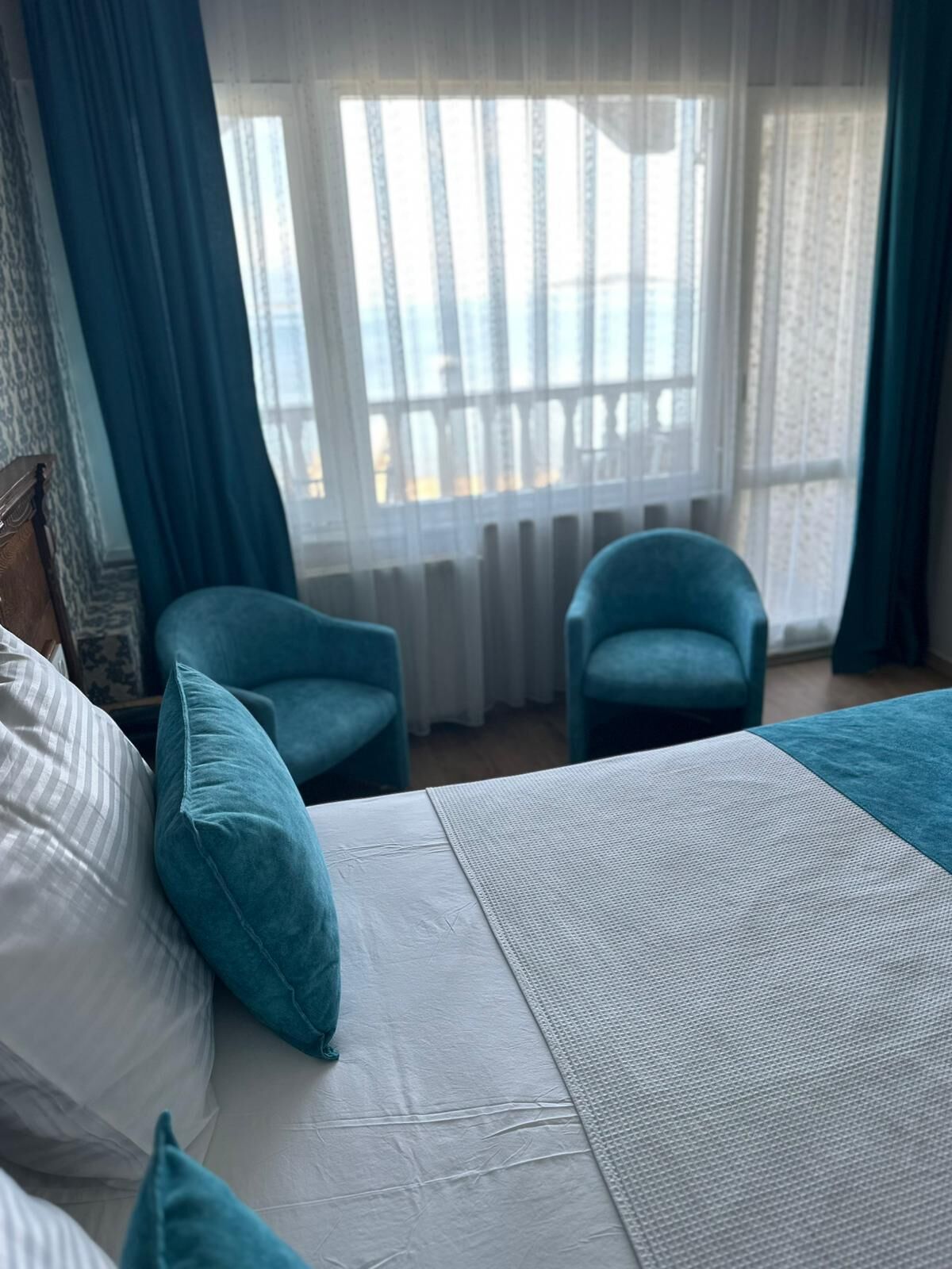 A hotel room with a bed, two chairs, a view of the sea through a window with curtains, and a wooden headboard.