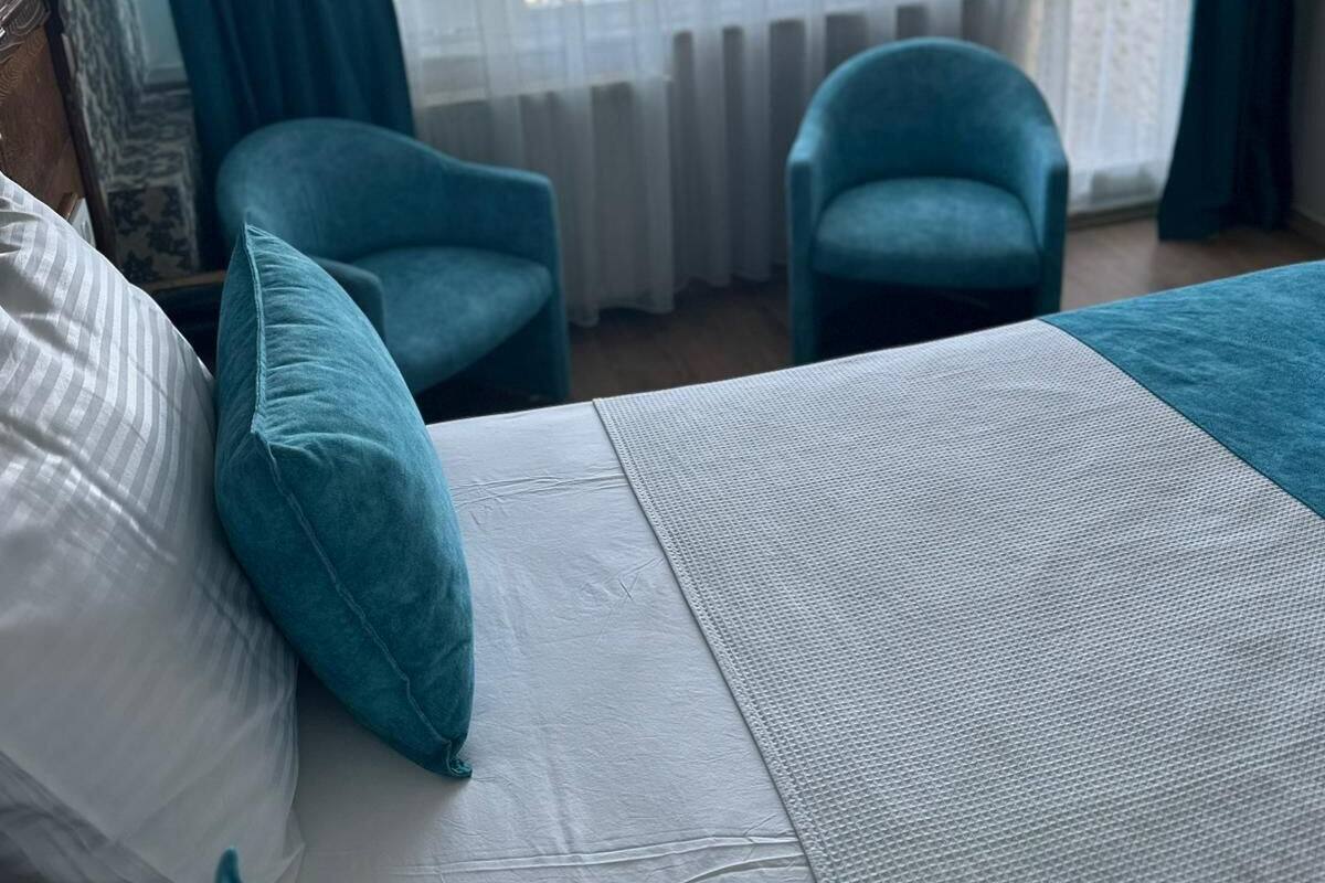 A hotel room with a bed, two chairs, a view of the sea through a window with curtains, and a wooden headboard.