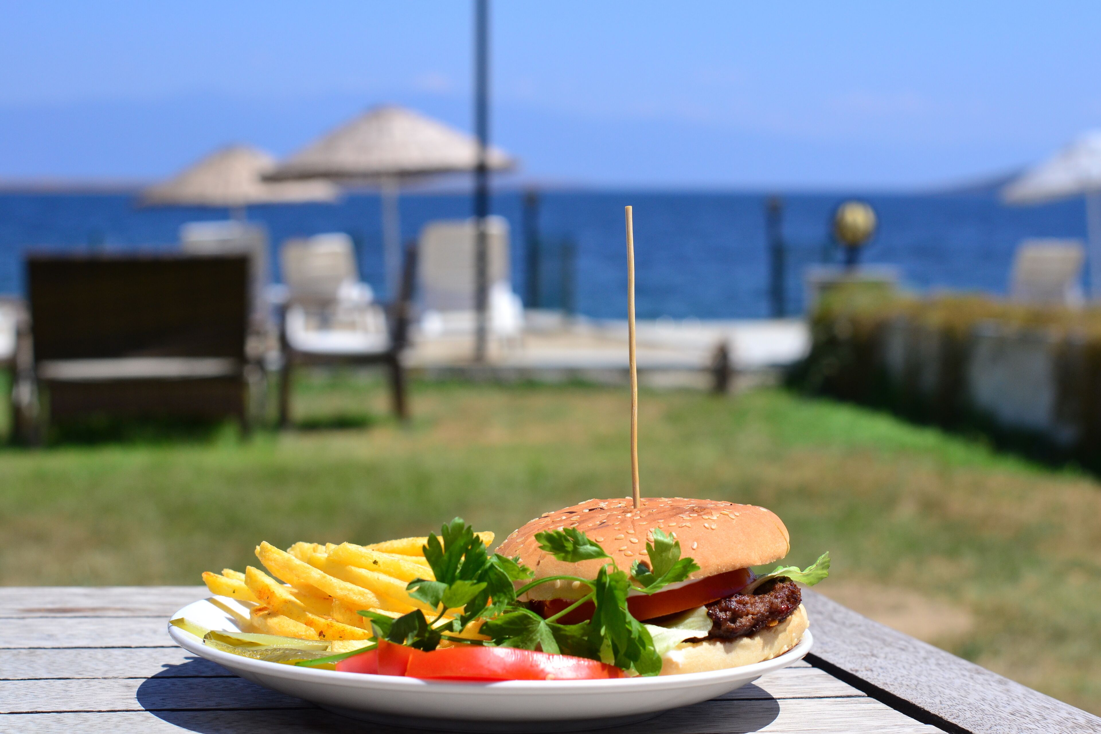 Un plato con una hamburguesa, papas fritas y verduras sobre una mesa de madera, con vista al mar y sombrillas de playa al fondo.