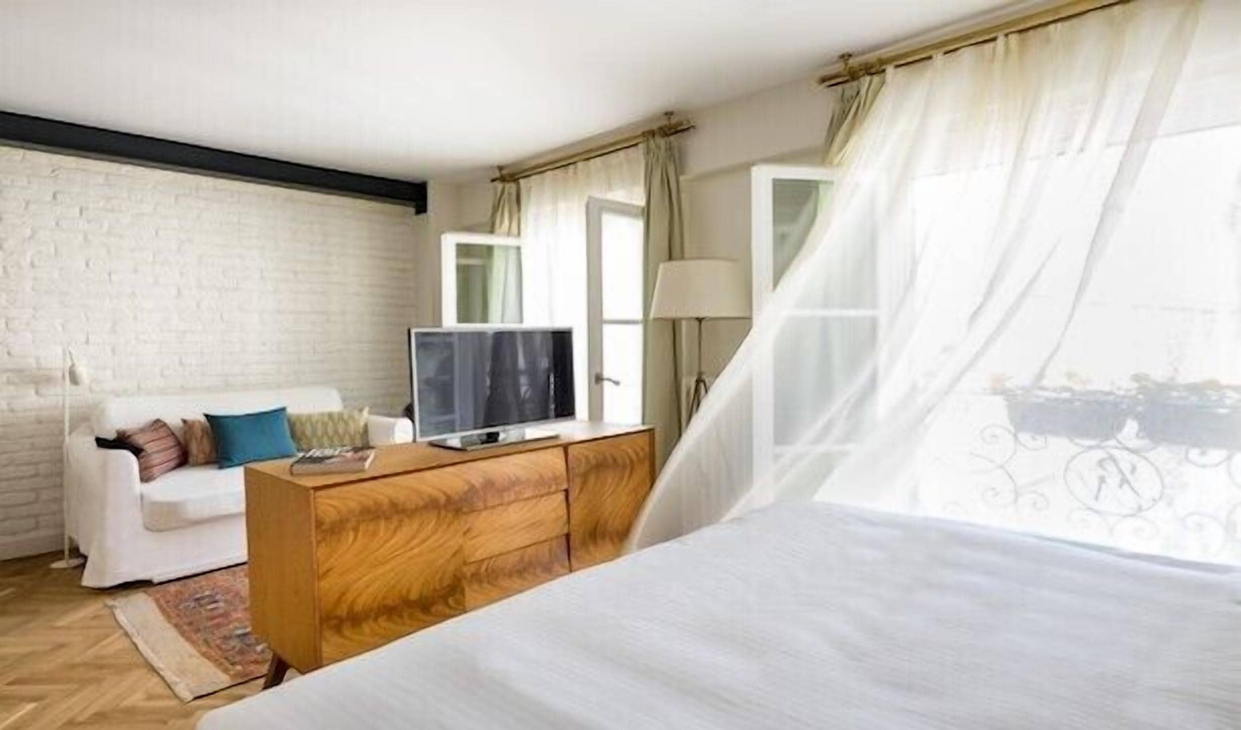 Une chambre à coucher avec un lit, un téléviseur posé sur une commode en bois, un canapé blanc et une fenêtre avec des rideaux.