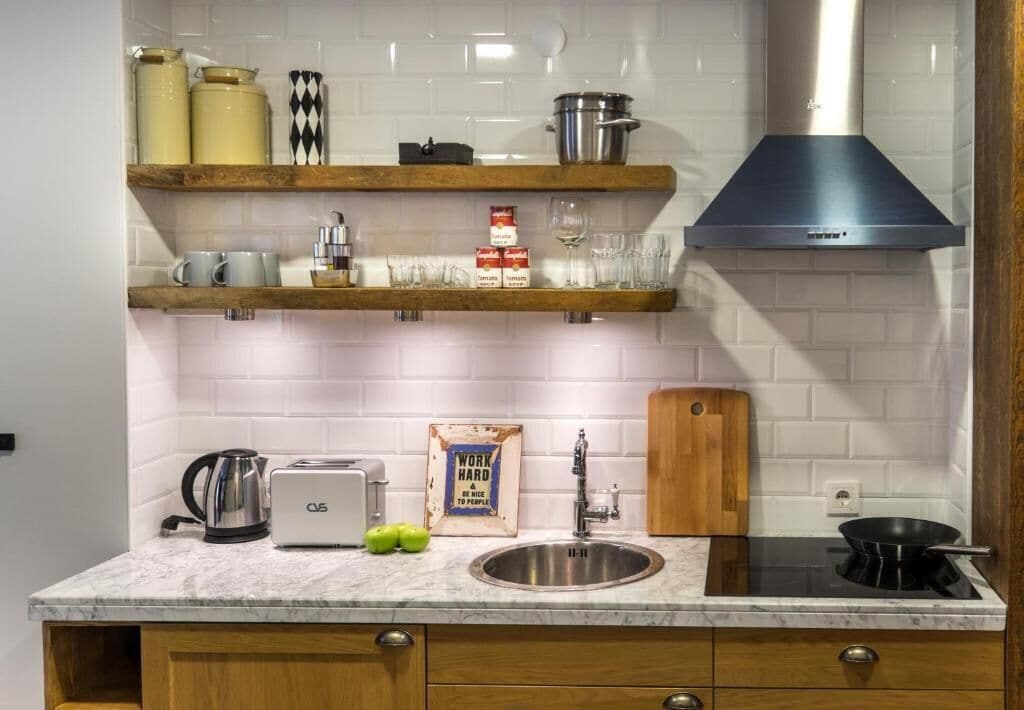 Una cocina con baldosas blancas tipo metro, estantes de madera, un lavamanos de acero inoxidable, un tostador, un hervidor y una estufa negra.
