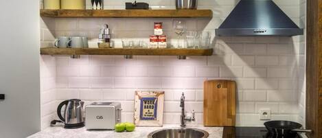 Una cocina con baldosas blancas tipo metro, estantes de madera, un lavamanos de acero inoxidable, un tostador, un hervidor y una estufa negra.