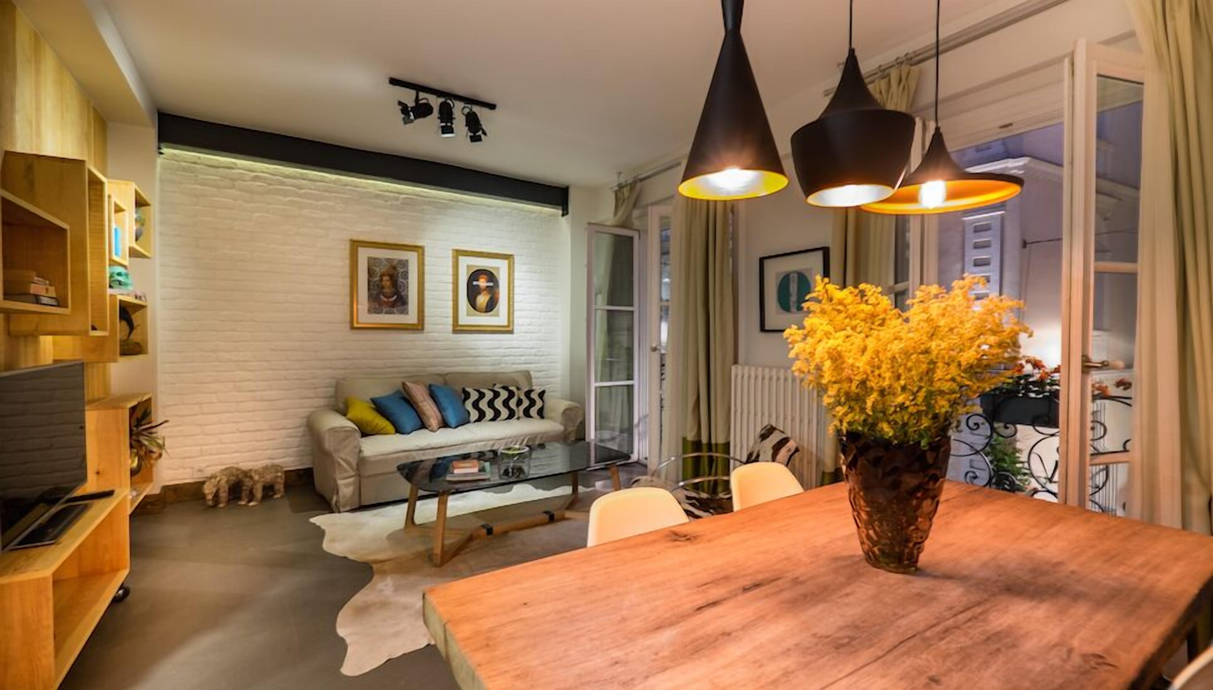 A cozy living room with a sofa, coffee table, and a wooden dining table. There are pendant lights above the table and a vase with flowers on the table.