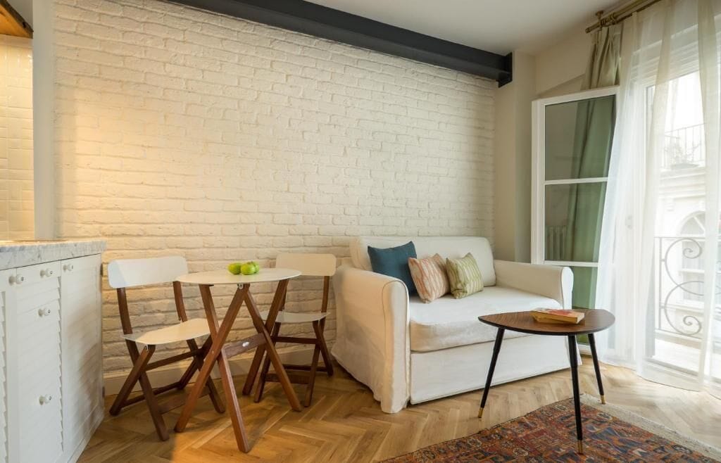 A cozy living area with a white sofa, a small round table, and a dining set against a brick wall.