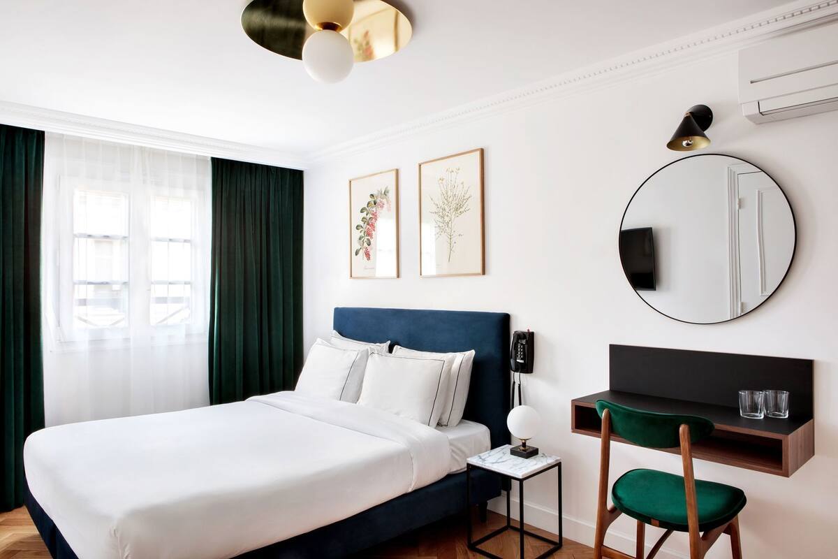 A hotel room with a bed, a desk, a chair, a round mirror, and two framed botanical prints on the wall.