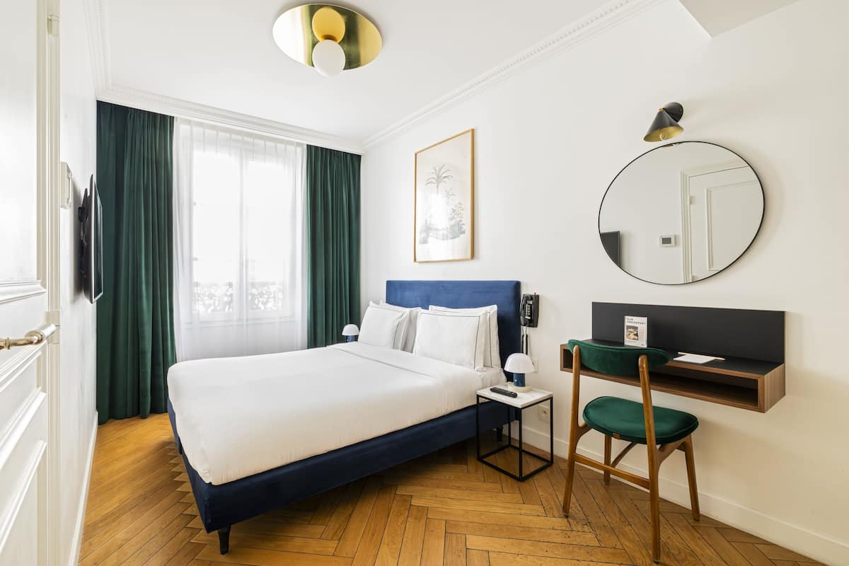 A hotel room with a bed, a desk, a chair, a round mirror, and a painting on the wall.