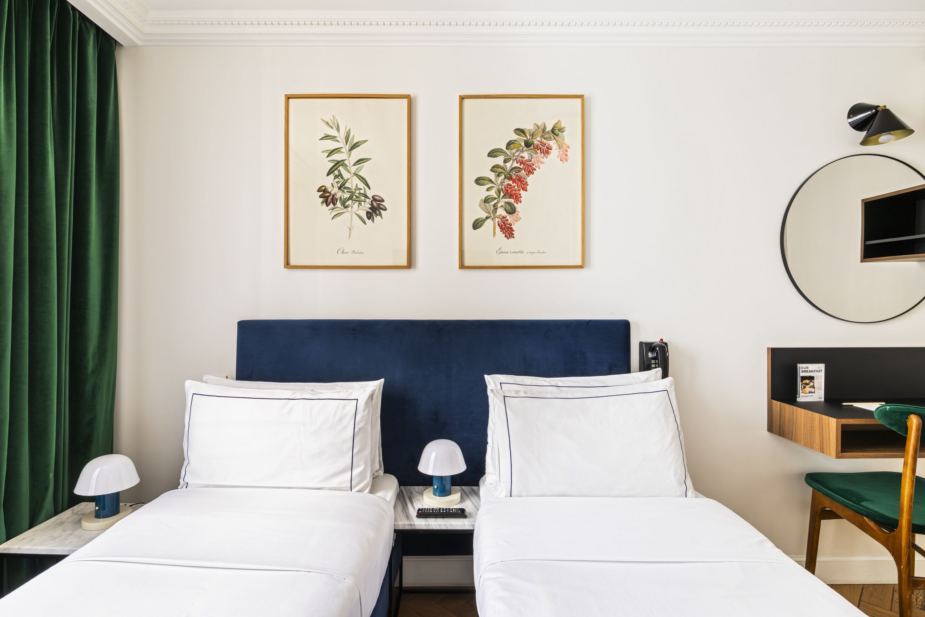 Ein Hotelzimmer mit zwei Betten, einem Nachttisch mit Lampe, einem Schreibtisch mit Stuhl und zwei gerahmten Pflanzenbildern an der Wand.